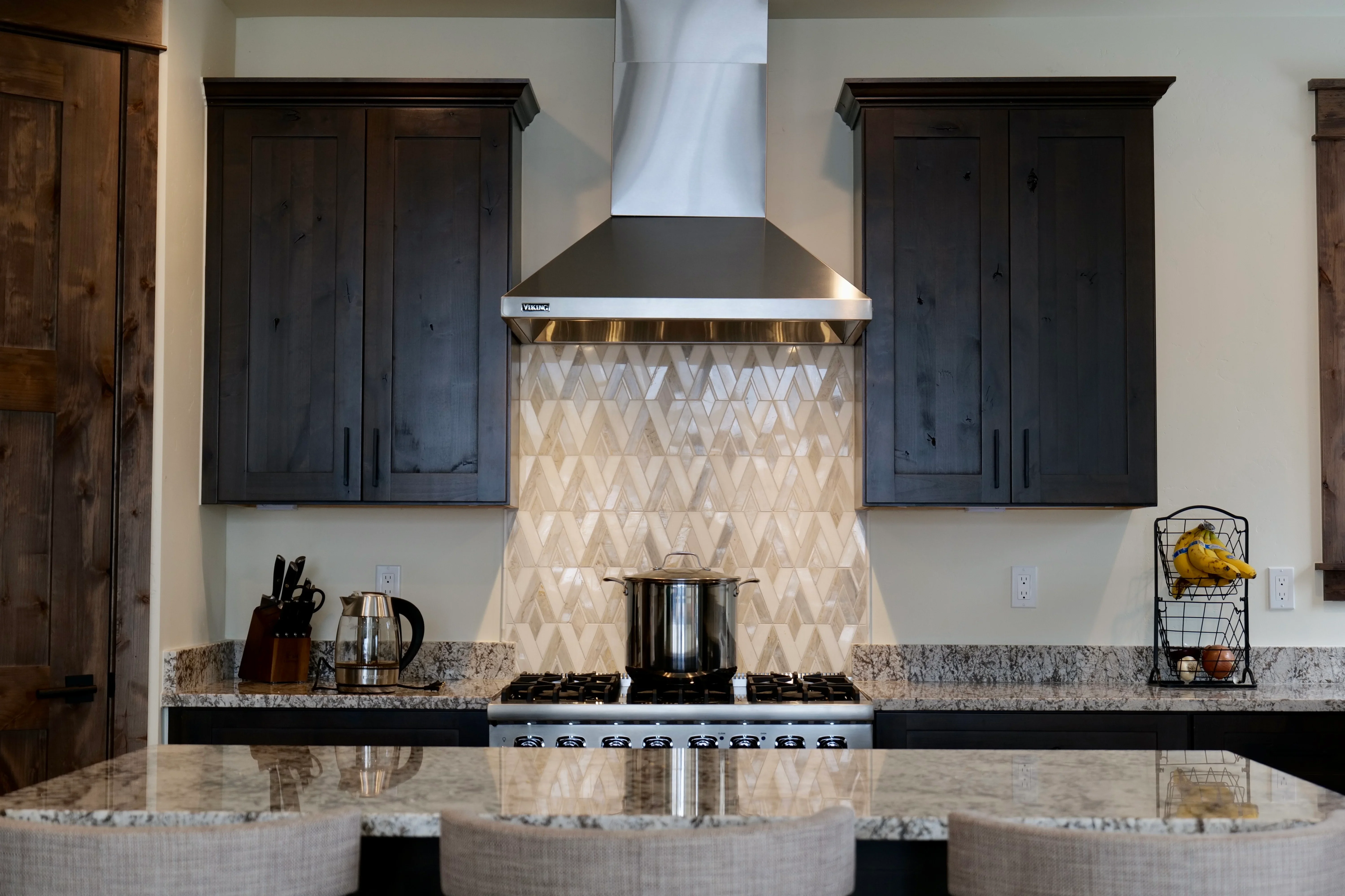 Dark wood cabinets, granite counters, geometric tile backsplash, gas range, stainless hood vent