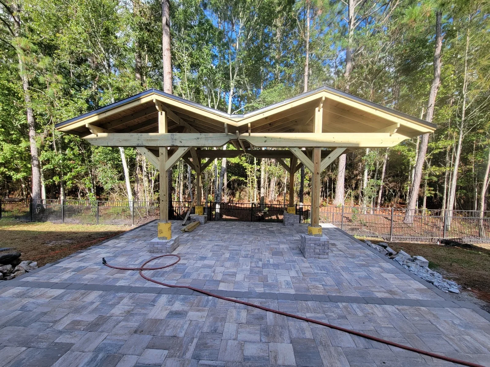 Timber frame pavilion with dual gable roofs and gray paver patio in wooded area