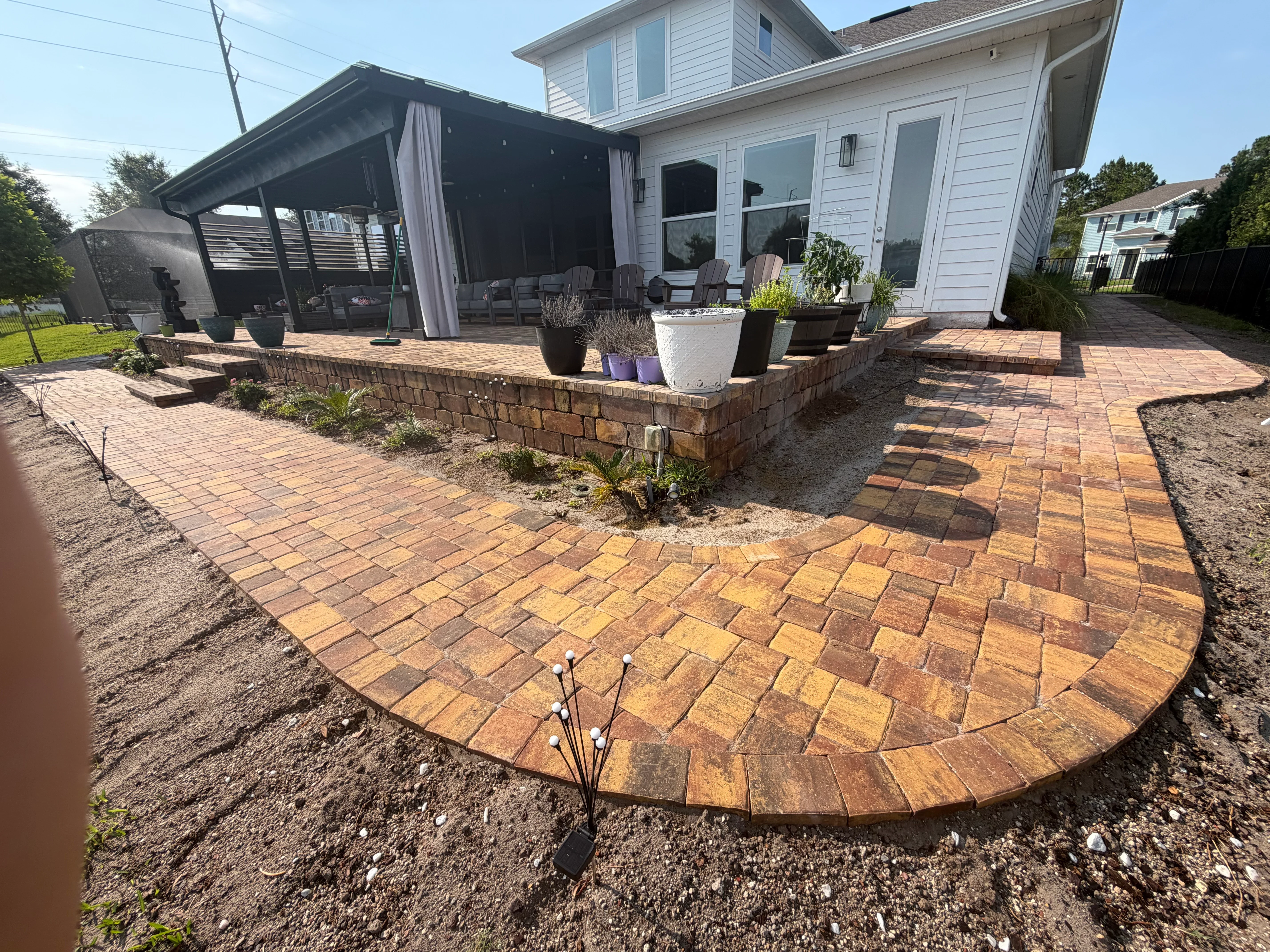 Curved brick paver patio with stone retaining wall and raised planter bed beside home