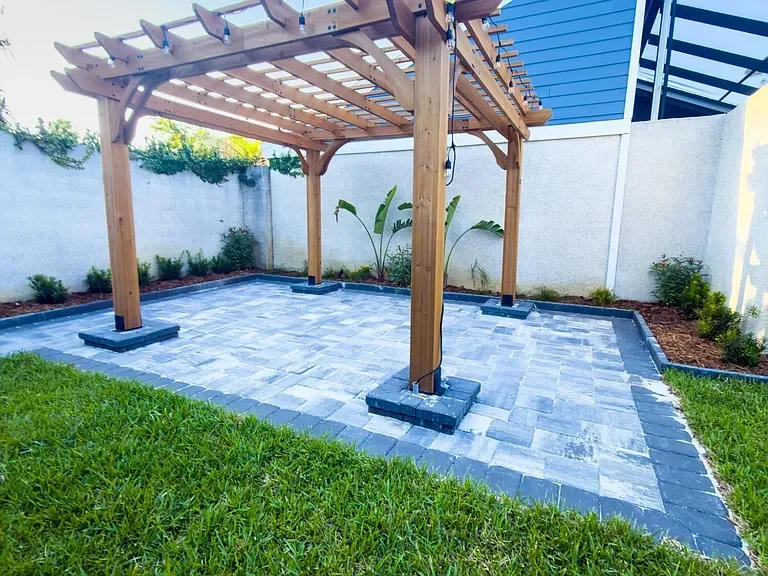 Wooden pergola with shade panels over blue-gray paver patio with concrete footings and landscaping
